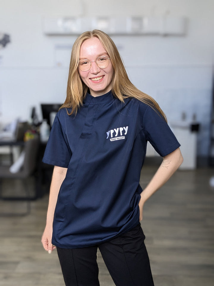 A person wearing a navy chef coat with a logo on the chest, standing in a kitchen environment.