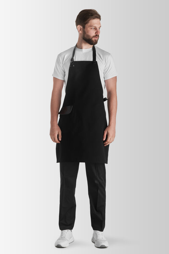 A person wearing a black Jackson canvas apron with an adjustable neck strap and a front pocket, paired with a white t-shirt and black pants.