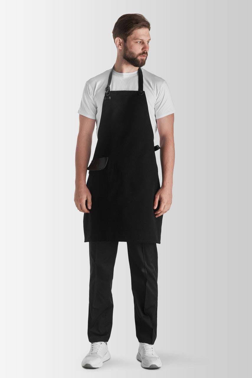 A person wearing a black Jackson canvas apron with an adjustable neck strap and a front pocket, paired with a white t-shirt and black pants.
