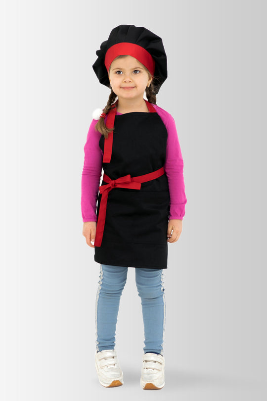 A child model wearing a black children's apron with a red tie and a matching toque, standing against a white background.