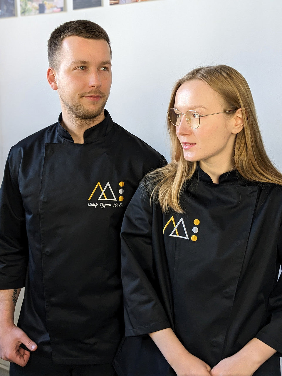 A man and a woman wearing black chef coats with a white logo, standing in a kitchen environment.