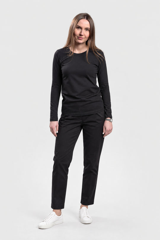 A woman modeling a black long-sleeve crewneck top and black pants with white sneakers, standing against a light gray studio background.