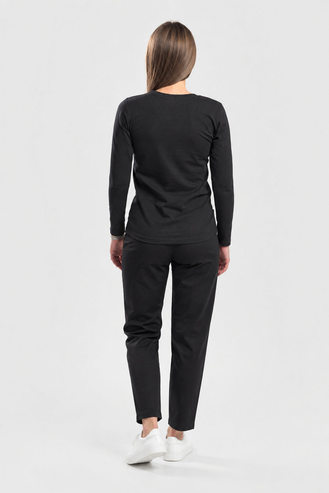 A woman viewed from behind wearing a black long-sleeve top and black pants with white sneakers, standing against a light gray studio background.