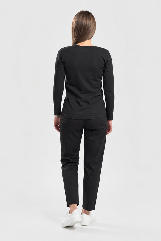 A woman viewed from behind wearing a black long-sleeve top and black pants with white sneakers, standing against a light gray studio background.