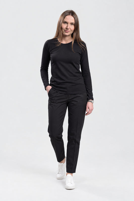 A woman modeling a black long-sleeve crewneck top and black trousers, paired with white sneakers, against a light gray studio background.