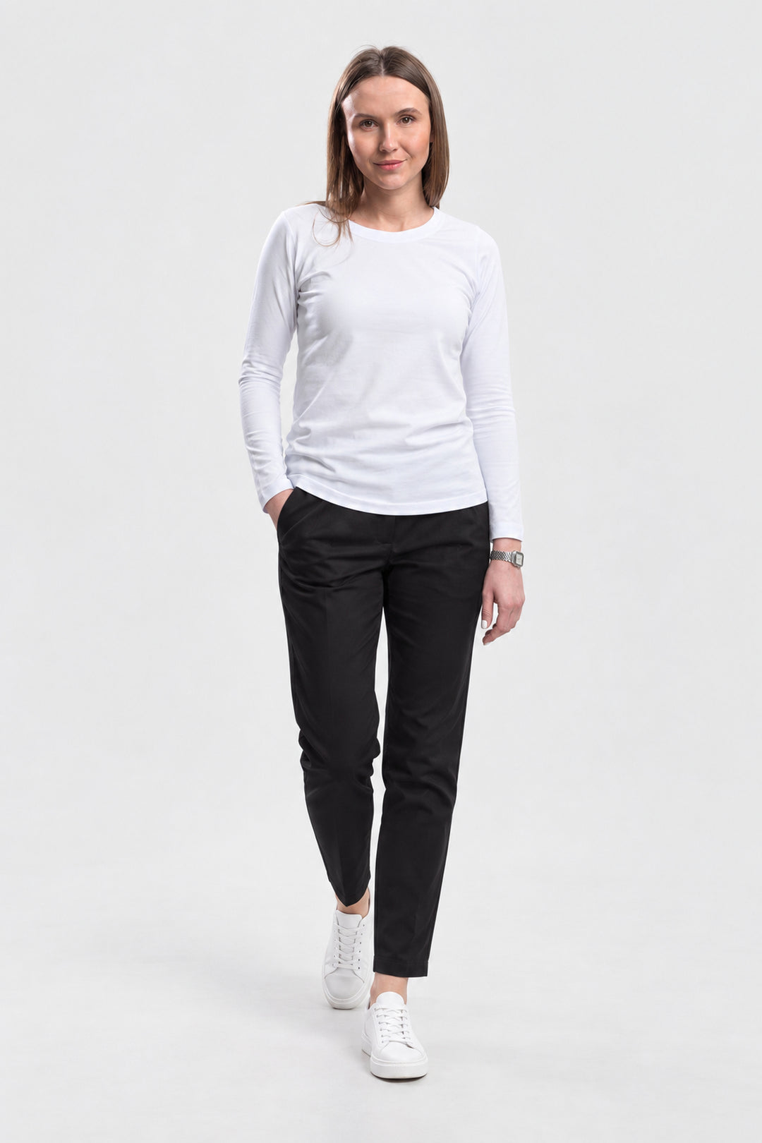 A woman modeling a white long-sleeve crewneck shirt, black slim-fit trousers, and white sneakers against a plain light gray studio background.