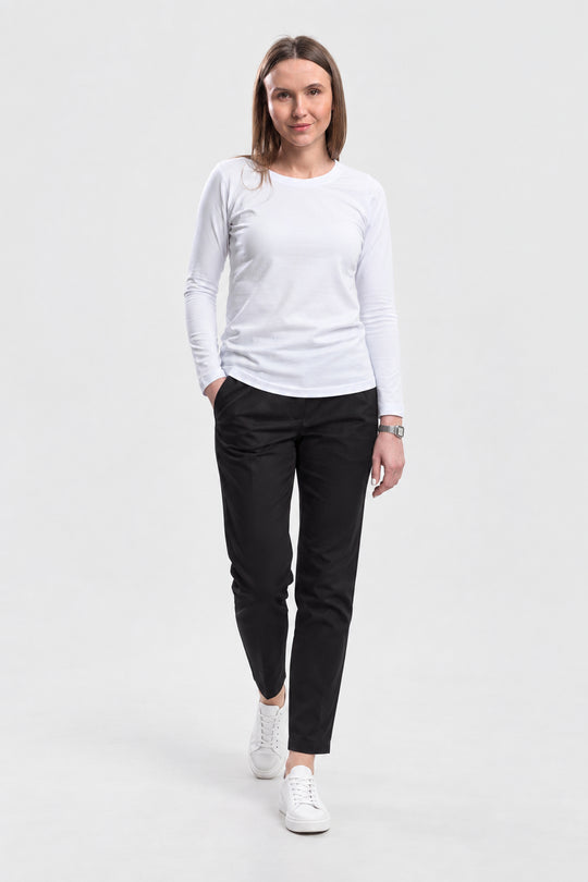 A woman modeling a white long-sleeve crewneck shirt, black slim-fit trousers, and white sneakers against a plain light gray studio background.