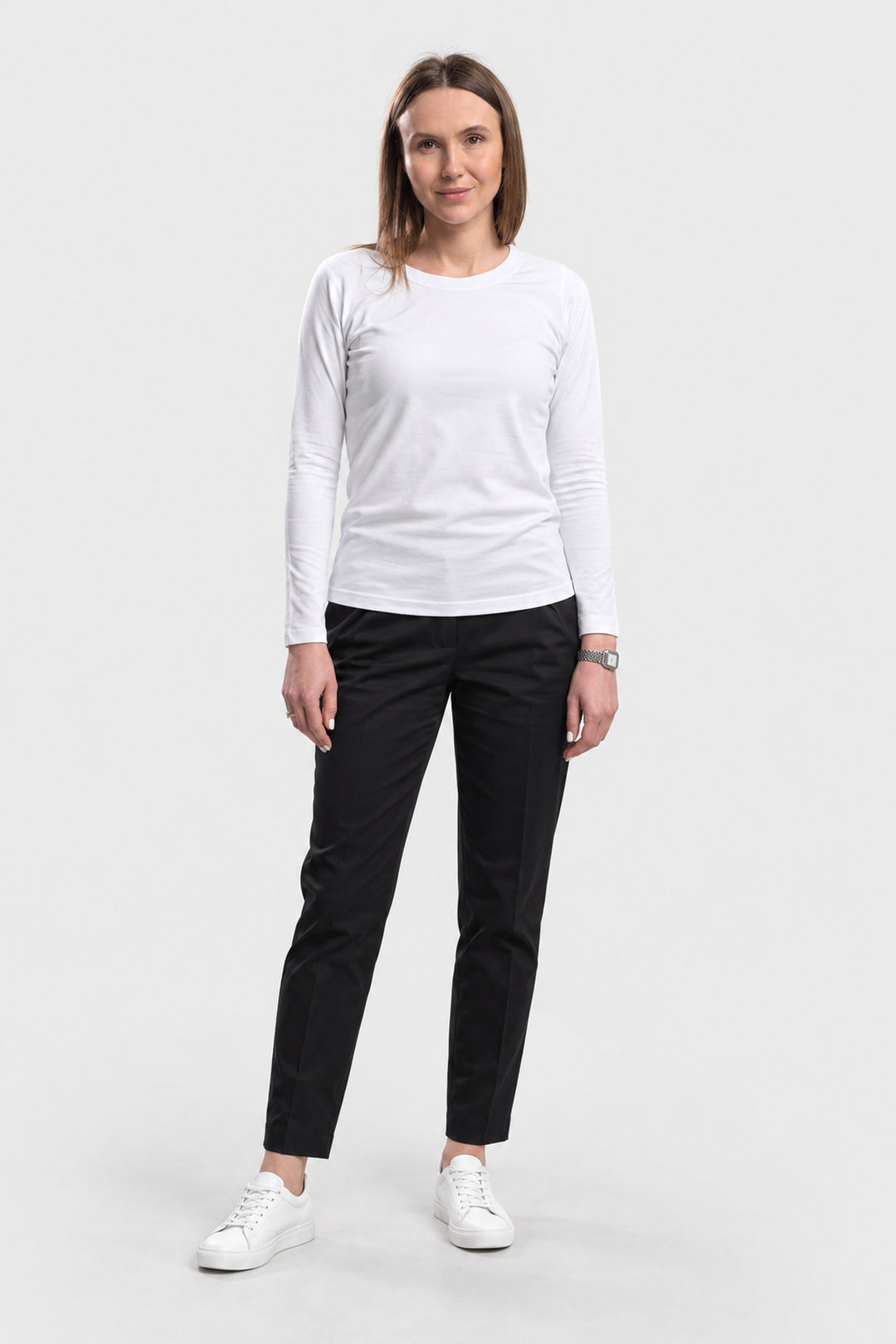 A woman modeling a white long-sleeve crewneck shirt and black slim-fit pants with white sneakers, standing against a light gray studio background.