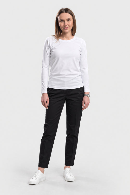 A woman modeling a white long-sleeve crewneck shirt and black slim-fit pants with white sneakers, standing against a light gray studio background.