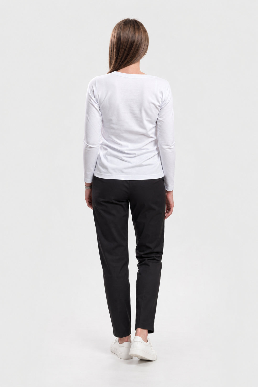 A woman viewed from behind wearing a white long-sleeve top, black pants, and white sneakers against a plain light gray studio background.