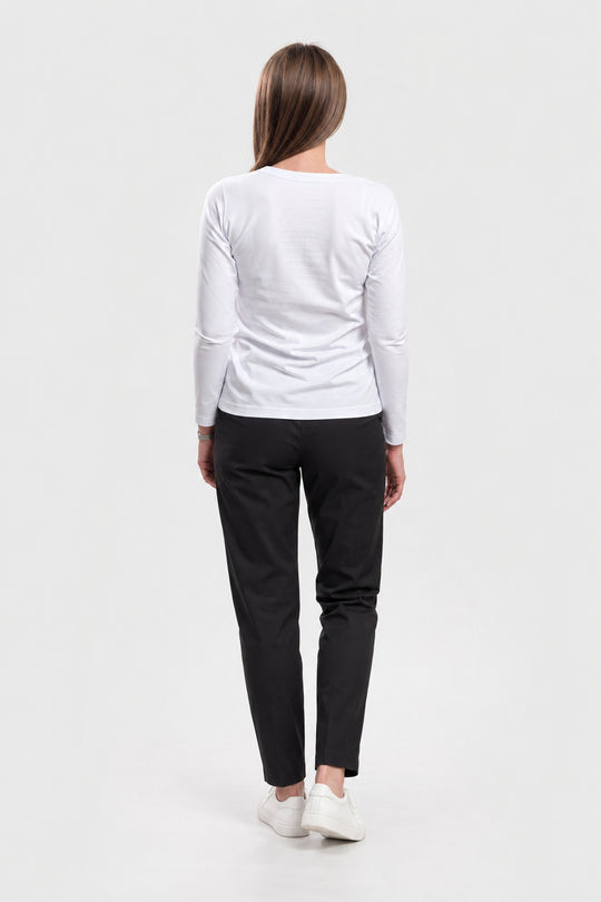 A woman viewed from behind wearing a white long-sleeve top, black pants, and white sneakers against a plain light gray studio background.