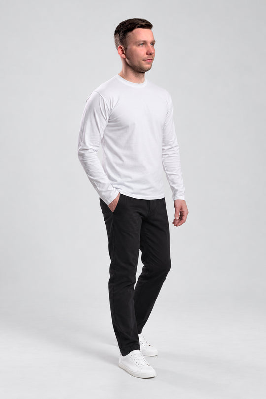 A man wearing a white long-sleeve crewneck shirt, black trousers, and white sneakers, standing in profile against a light gray studio background.