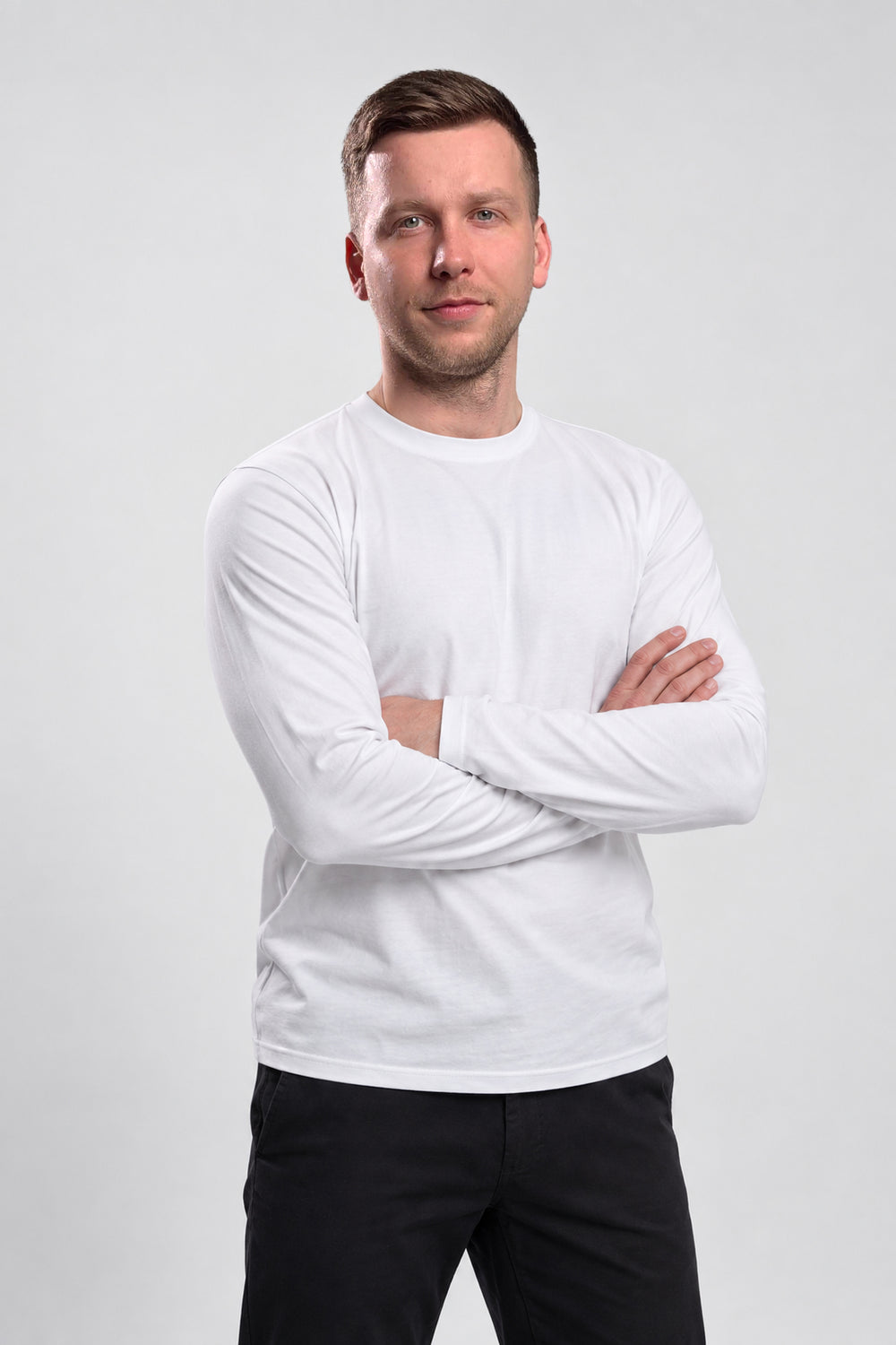 A man wearing a plain white long-sleeve crewneck shirt and black pants, standing with arms crossed against a light gray studio background.