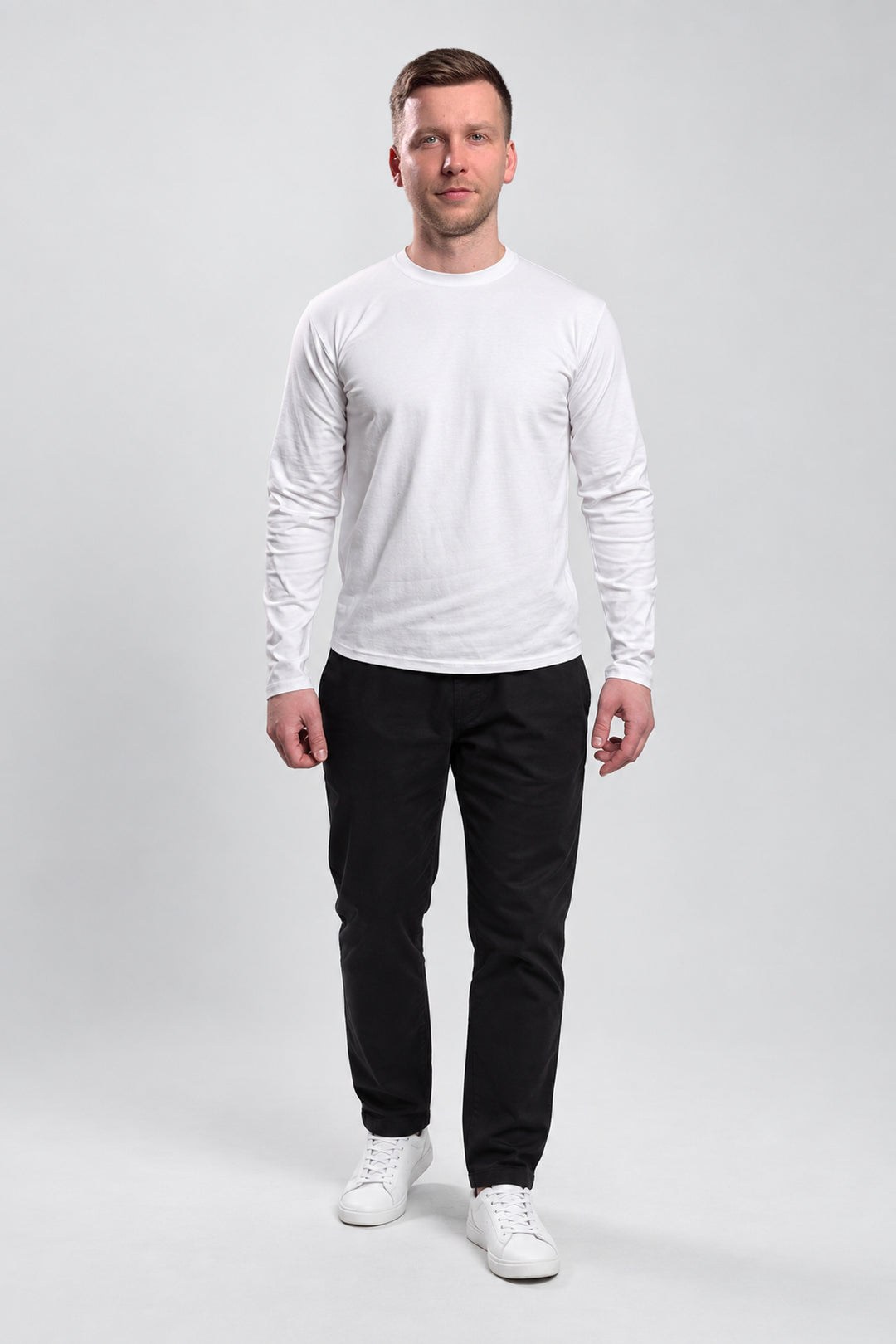 A man modeling a white long-sleeve crewneck shirt paired with black pants and white sneakers, standing against a light gray studio background.