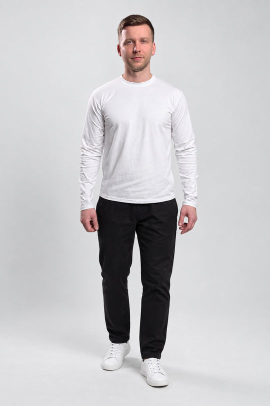 A man modeling a white long-sleeve crewneck shirt paired with black pants and white sneakers, standing against a light gray studio background.