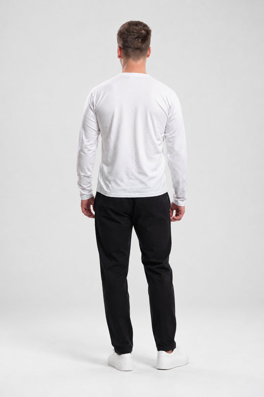 A man viewed from behind wearing a white long-sleeve crewneck shirt, black trousers, and white sneakers, standing against a light gray studio background.