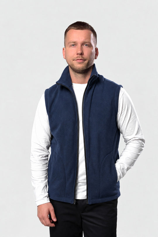 A man wearing a navy blue fleece vest over a white long-sleeve shirt and black pants, standing against a light gray studio background.