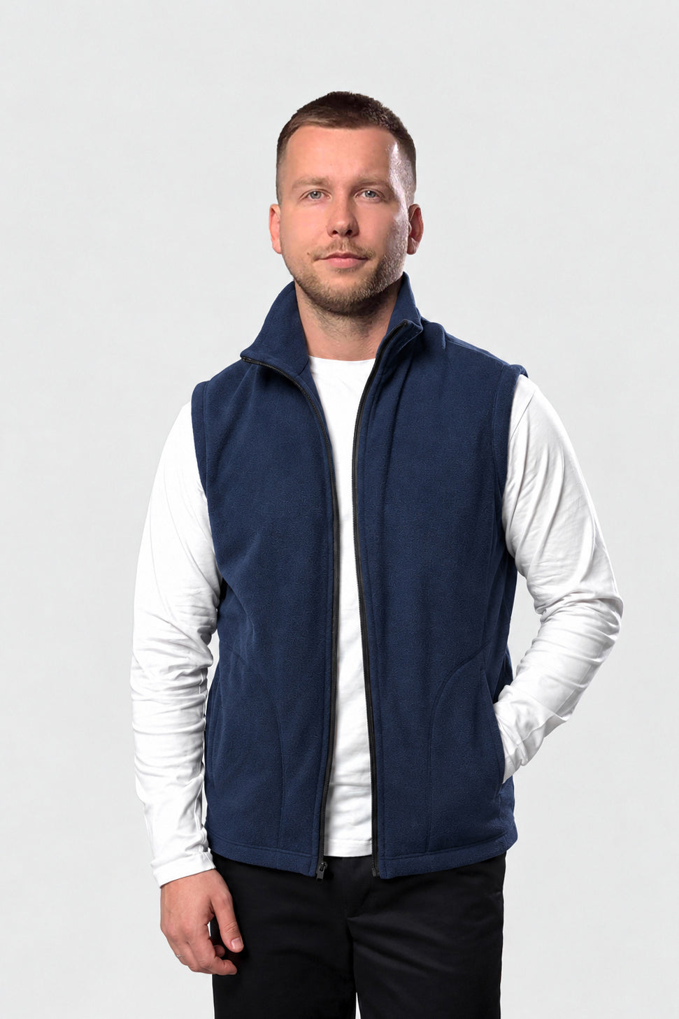 A man wearing a navy blue fleece vest over a white long-sleeve shirt and black pants, standing against a light gray studio background.