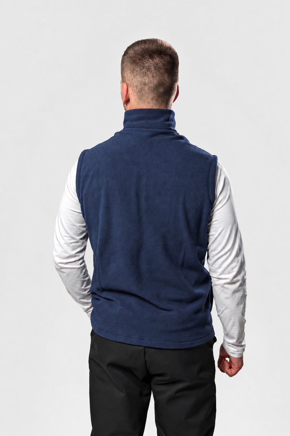 A man viewed from behind wearing a navy blue sleeveless fleece vest over a white long-sleeve shirt and black pants, against a light gray studio background.