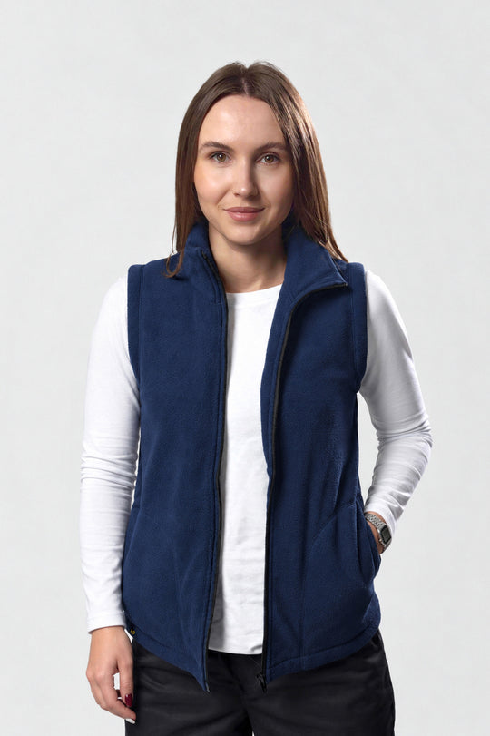 A woman wearing a navy blue fleece vest over a white long-sleeve shirt and black pants, standing against a light gray studio background.