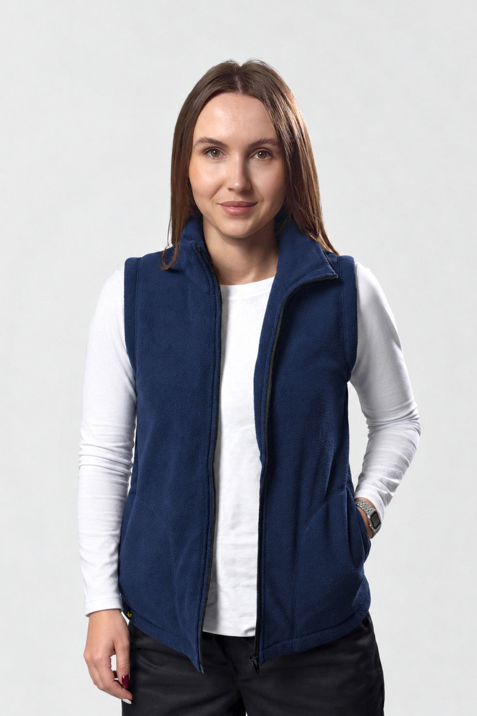 A woman wearing a navy blue fleece vest over a white long-sleeve shirt and black pants, standing against a light gray studio background.