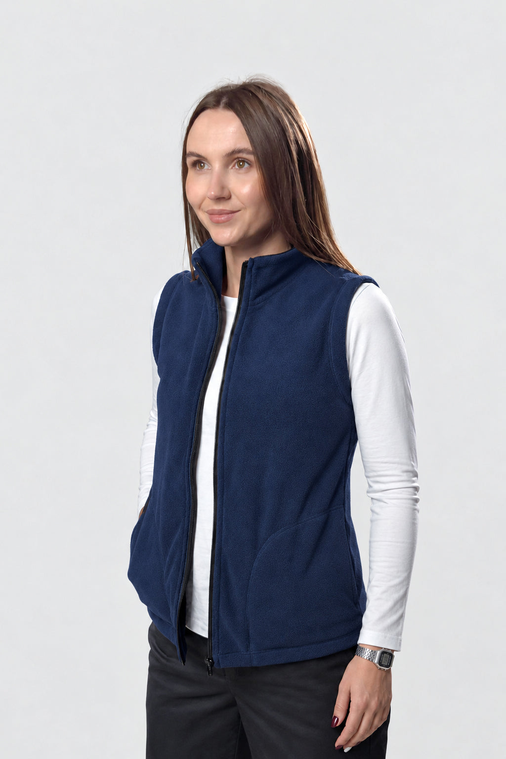 A woman modeling a navy blue zip-up fleece vest over a white long-sleeve shirt and dark pants, photographed against a light gray studio background.