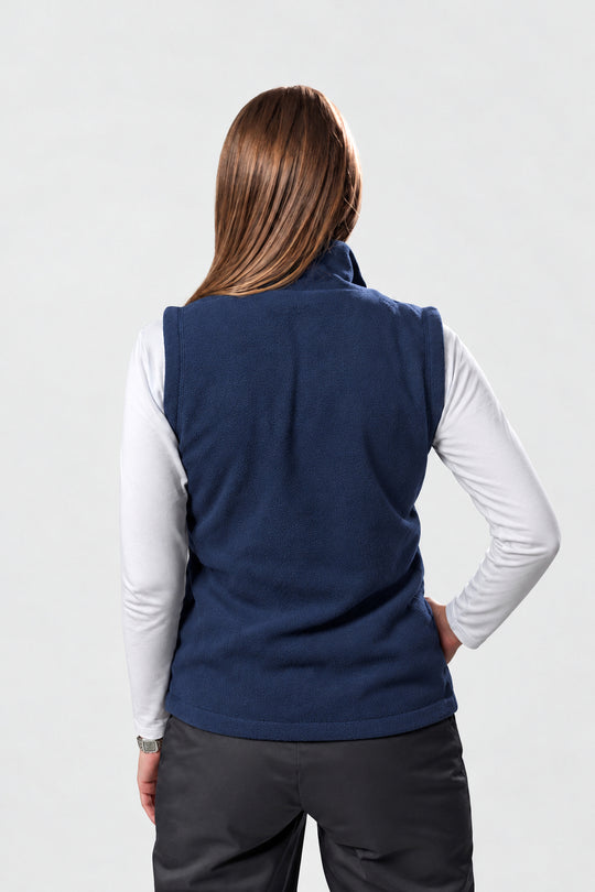 A woman viewed from the back wearing a navy blue sleeveless fleece vest over a white long-sleeve shirt and dark gray pants, against a light gray studio background.