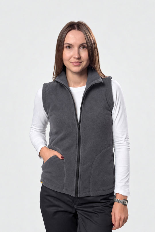 A woman modeling a dark gray zip-up fleece vest over a white long-sleeve shirt and black pants, photographed against a light gray studio background.