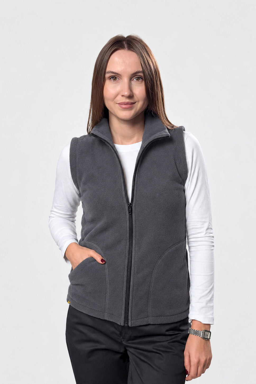 A woman modeling a dark gray zip-up fleece vest over a white long-sleeve shirt and black pants, photographed against a light gray studio background.