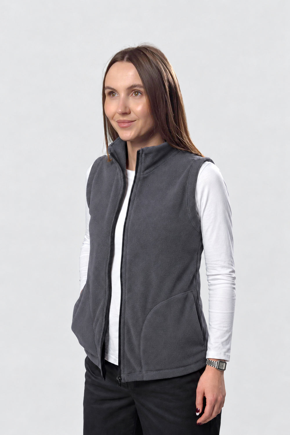 A woman wearing a dark gray zip-up fleece vest over a white long-sleeve shirt and black pants, photographed against a light gray studio background.