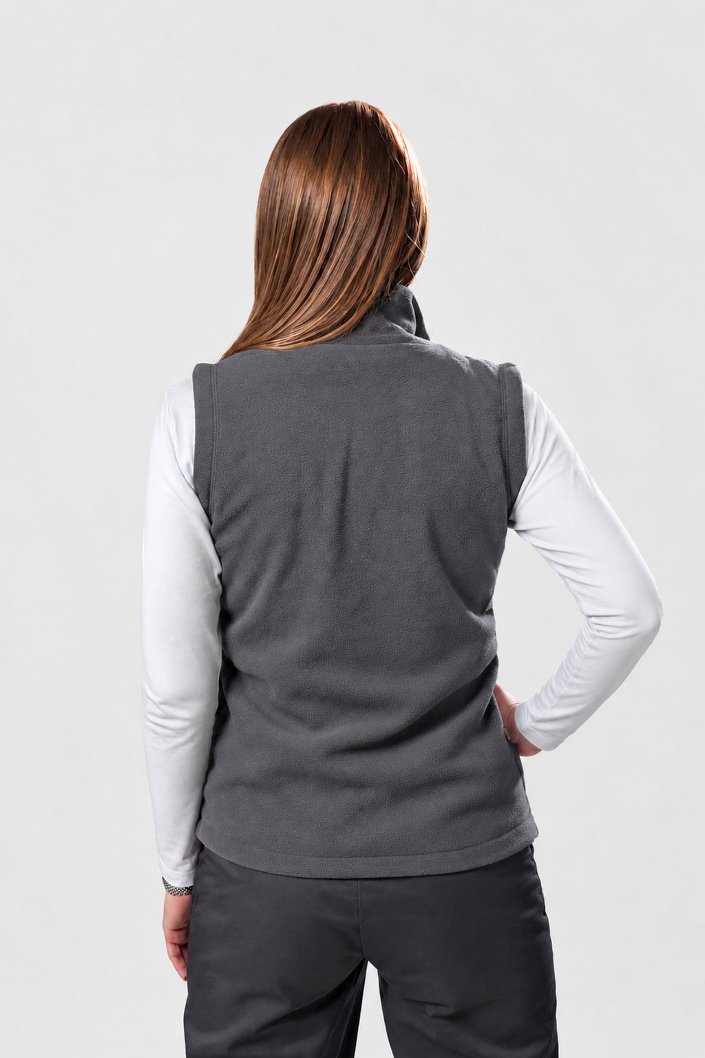 A woman shown from the back wearing a dark gray sleeveless fleece vest over a white long-sleeve shirt and dark gray pants, against a light gray studio background.
