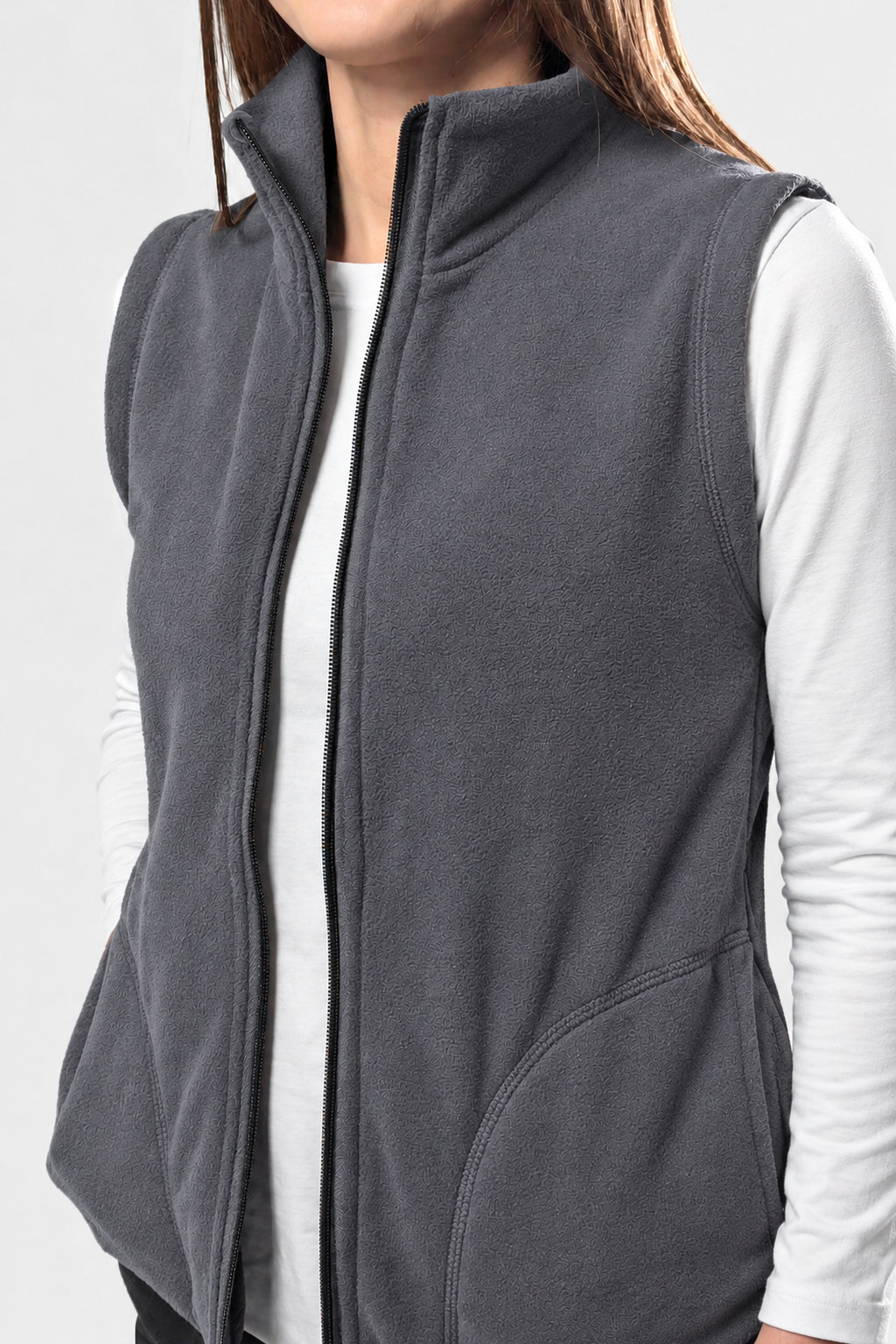 A close-up of a person wearing a dark gray fleece vest over a white long-sleeve shirt, cropped from the lower face to the waist against a light gray studio background.
