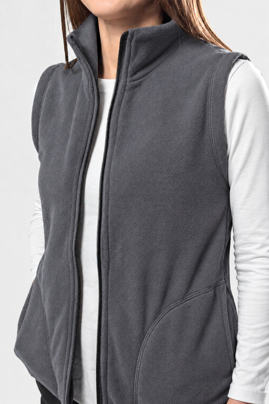 A close-up of a person wearing a dark gray fleece vest over a white long-sleeve shirt, cropped from the lower face to the waist against a light gray studio background.