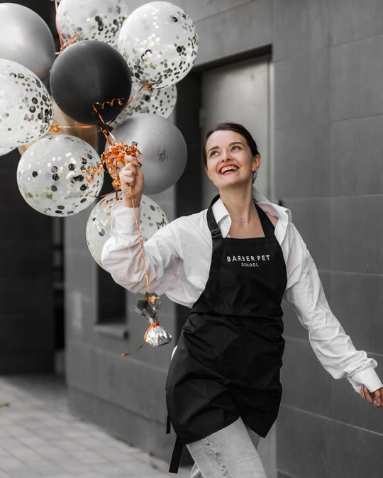 Woman wearing a black bib apron with embroidered logo while holding balloons outside a barber school