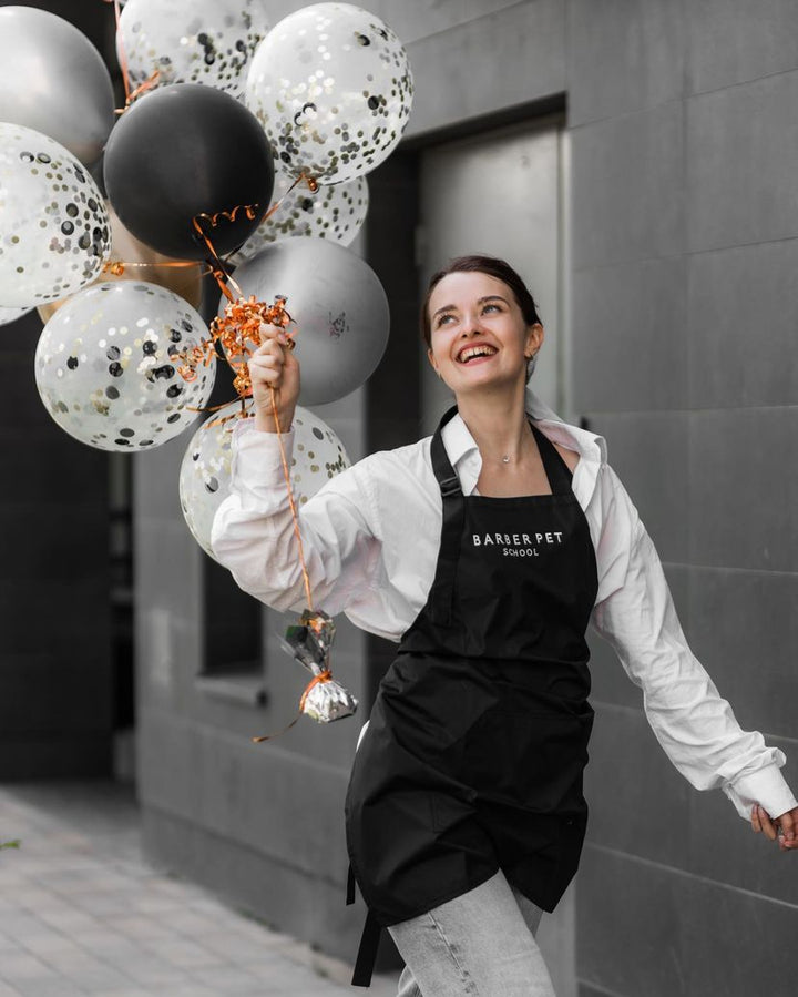 Woman wearing a black bib apron with embroidered logo while holding balloons outside a barber school