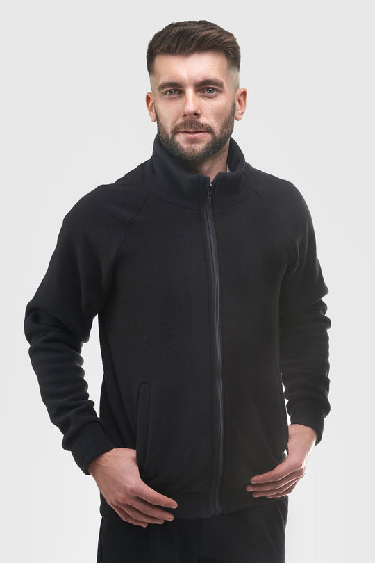 A man wearing a black zip-up fleece jacket with a stand-up collar, photographed against a solid light gray background.
