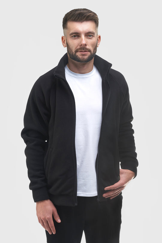 A man wearing a black zip-up fleece jacket over a white t-shirt and black pants, standing against a light gray studio background.