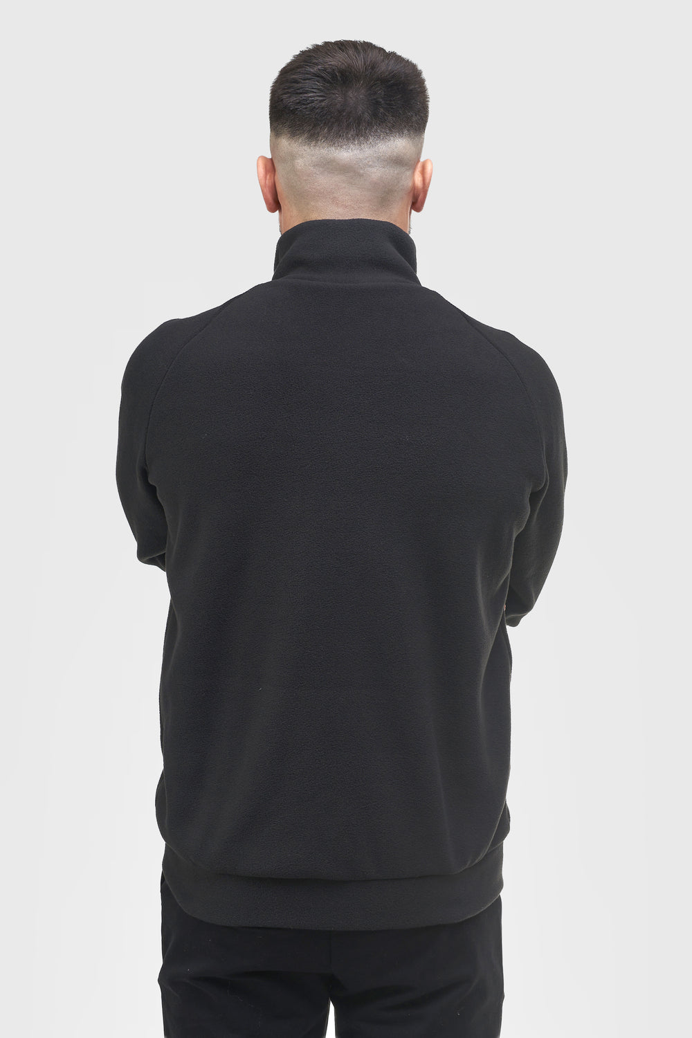 A man viewed from behind wearing a black mock-neck or high-collar long-sleeve top and black pants against a light gray studio background.