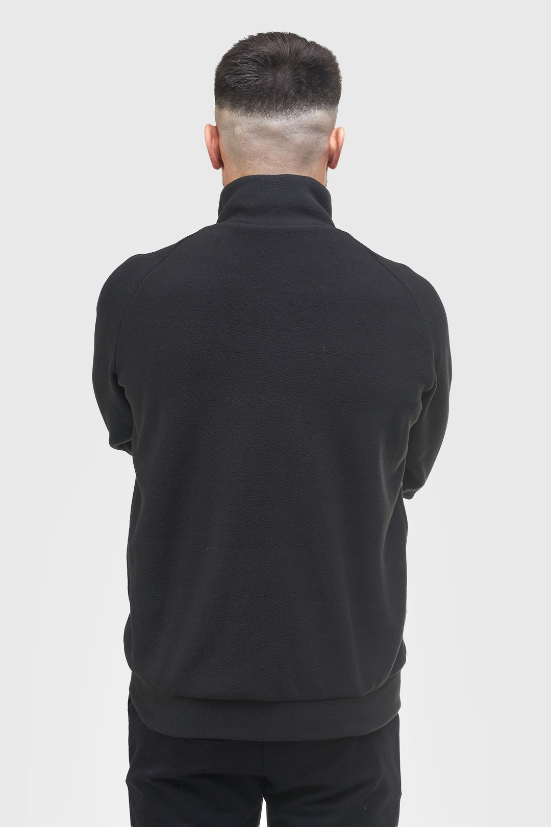 A man viewed from behind wearing a black mock-neck or high-collar long-sleeve top and black pants against a light gray studio background.