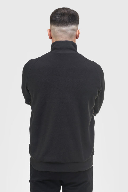 A man viewed from behind wearing a black mock-neck or high-collar long-sleeve top and black pants against a light gray studio background.