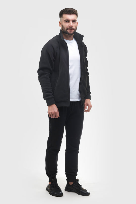 A man modeling a black zip-up fleece jacket over a white t-shirt, black jogger pants, and black sneakers, standing against a light gray studio background.