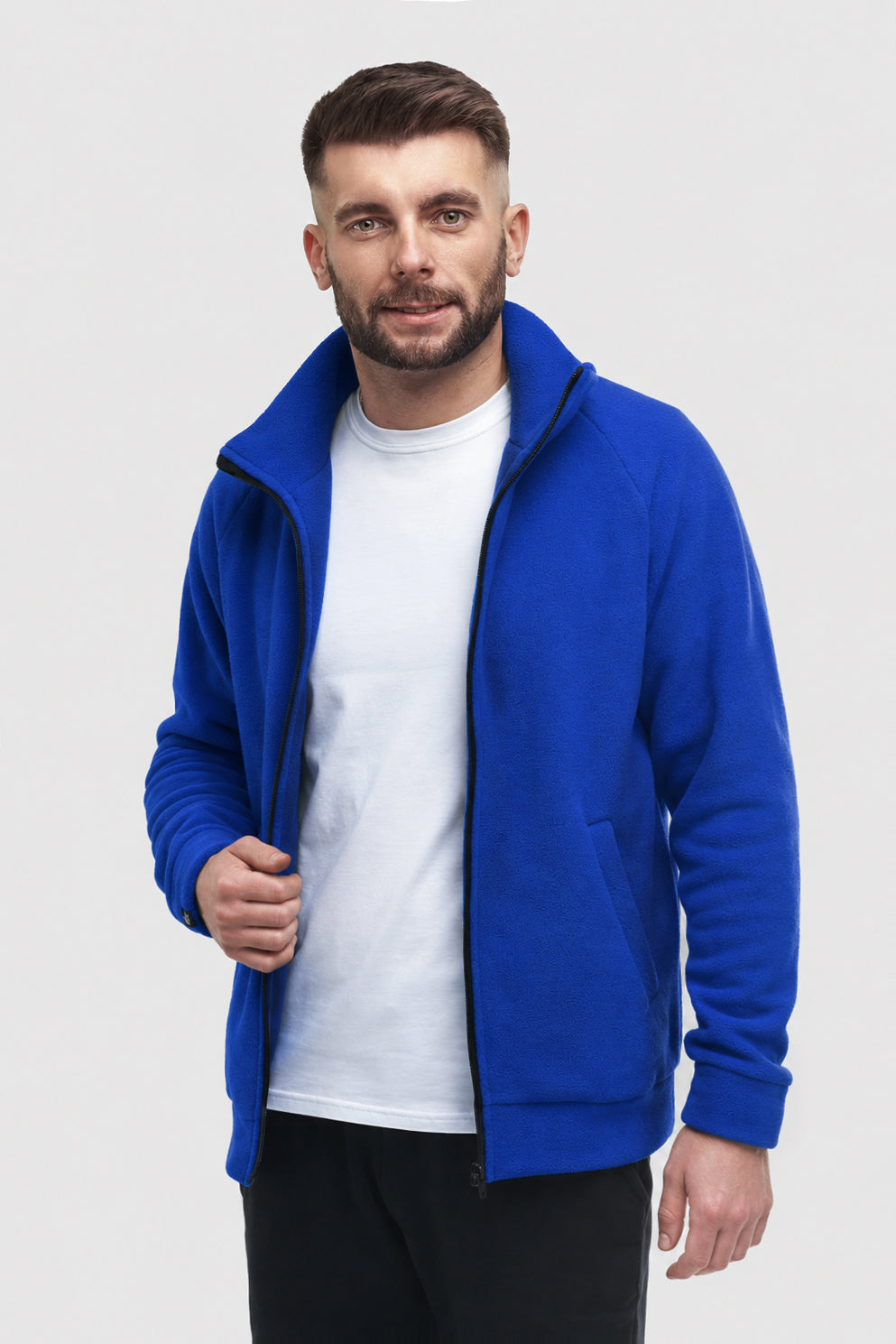 A man wearing a bright royal blue zip-up fleece jacket over a white t-shirt and black pants, standing against a light gray studio background.