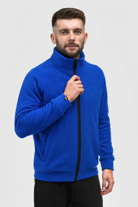 A man wearing a bright royal blue fleece zip-up jacket with a high collar, black zipper, and black pants, standing against a light gray studio background.