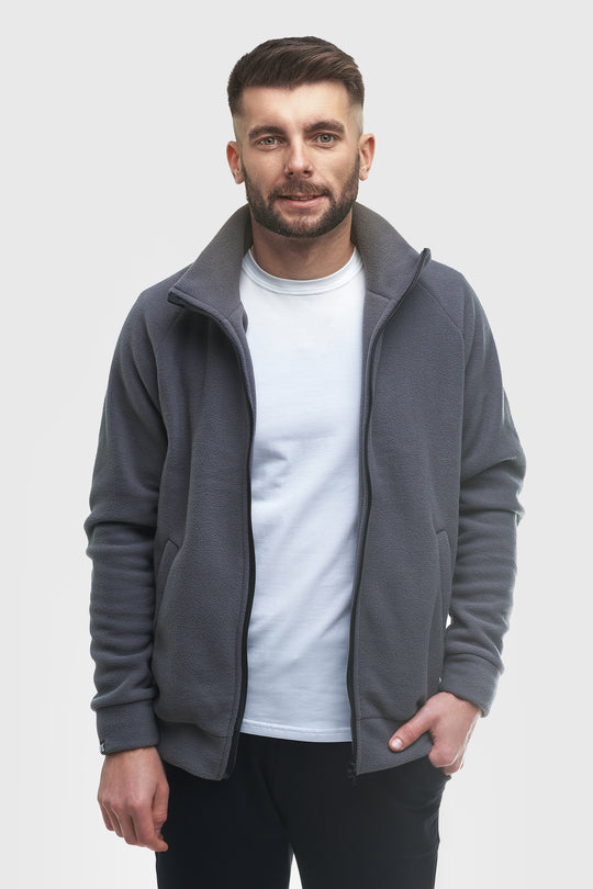A man wearing a dark gray zip-up fleece jacket over a white t-shirt and black pants, standing against a light gray studio background.