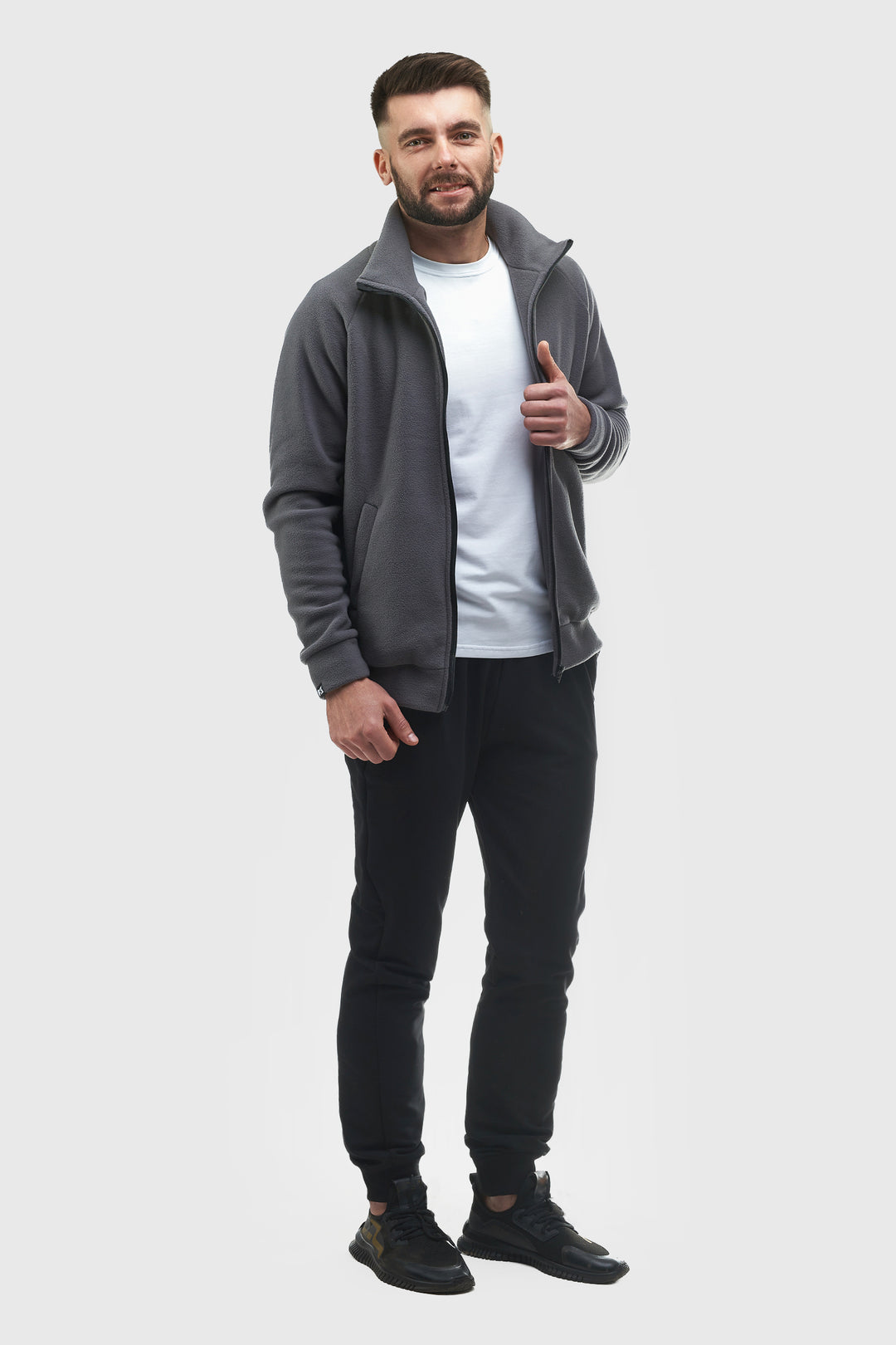 A man modeling a dark gray zip-up fleece jacket over a white t-shirt and black jogger pants, standing against a solid light gray background.