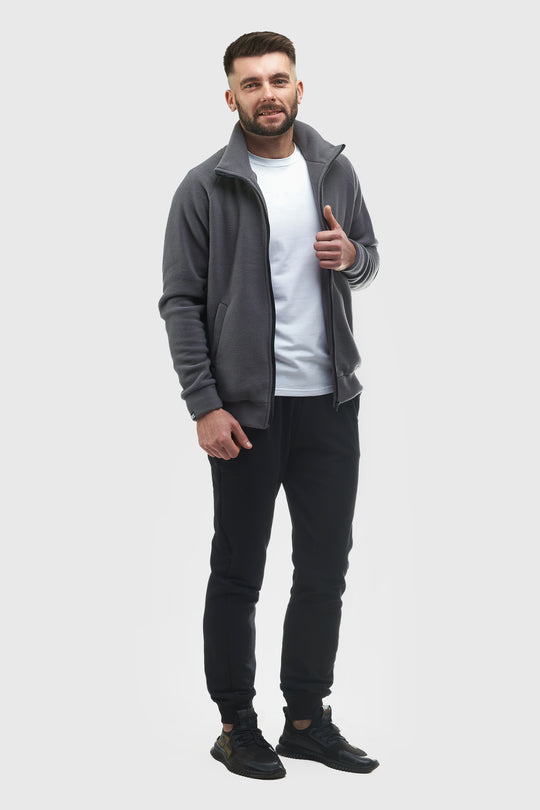 A man modeling a dark gray zip-up fleece jacket over a white t-shirt and black jogger pants, standing against a solid light gray background.