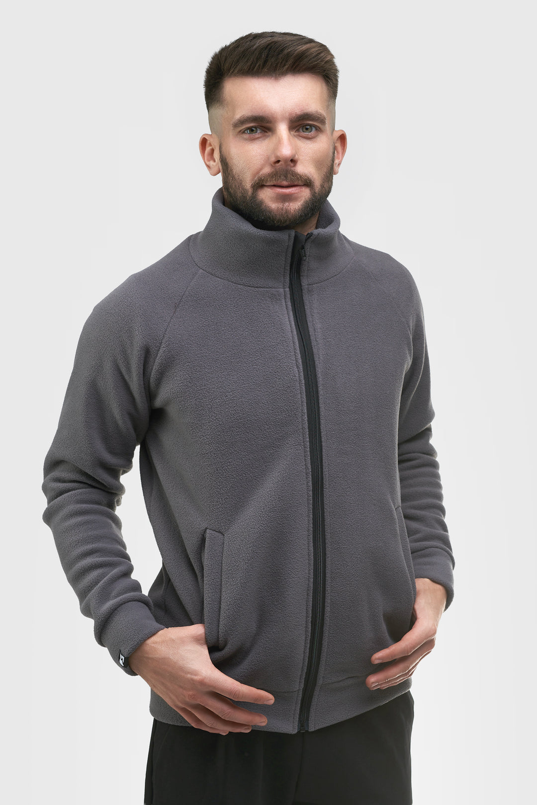 A man wearing a dark gray zip-up fleece jacket with a stand-up collar, shown from the waist up against a light gray studio background.