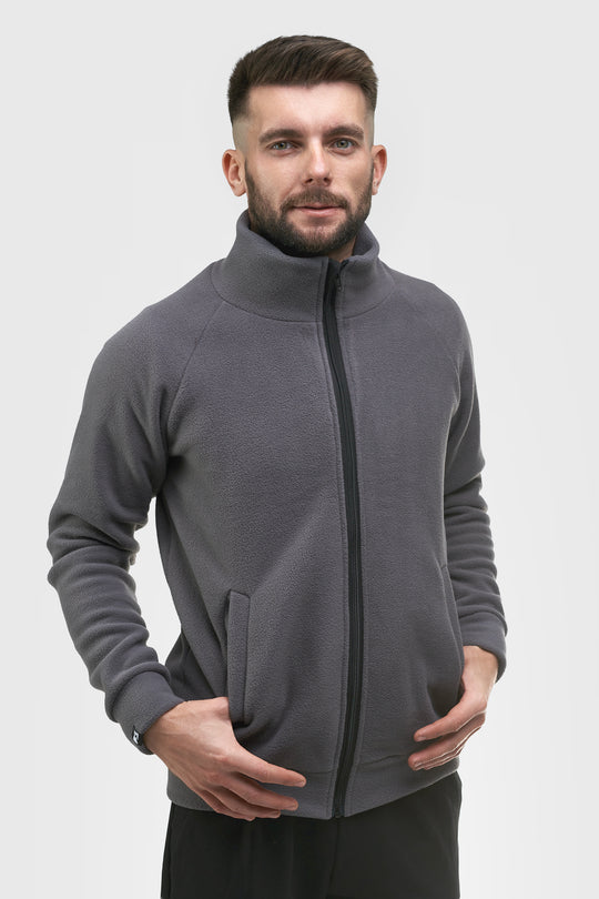 A man wearing a dark gray zip-up fleece jacket with a stand-up collar, shown from the waist up against a light gray studio background.