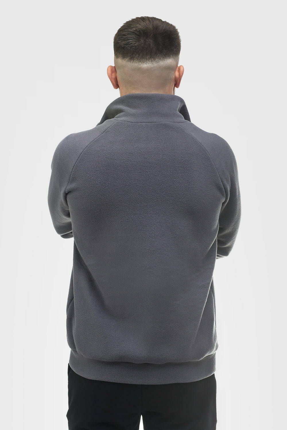 A man with a beard and short hair stands with arms crossed, wearing a dark gray zip-up fleece jacket over a white t-shirt and black pants, against a light gray studio background.