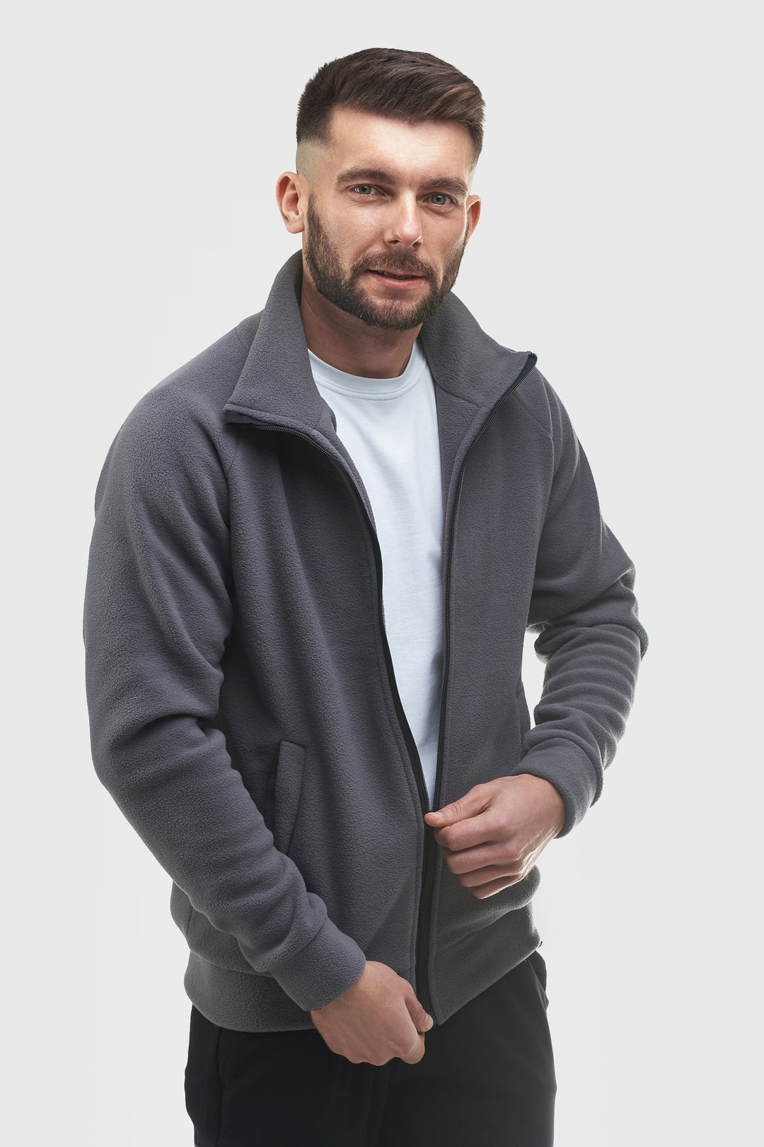 A man wearing a dark gray zip-up fleece jacket over a white t-shirt, standing against a light gray studio background.